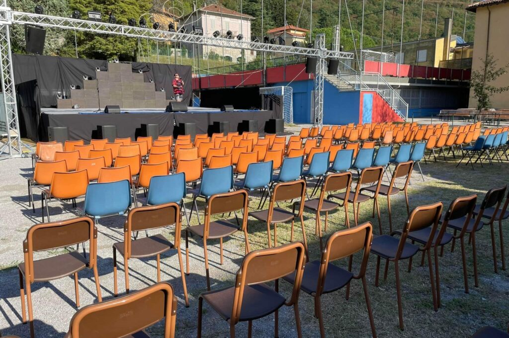 Palco all'aperto della Festa Rionale di Maggianico con file di sedie predisposte per il pubblico, impianto audio e luci per spettacoli ed eventi nell'area della festa.